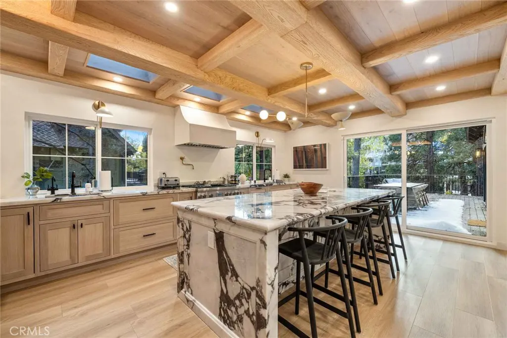 Bright kitchen with wood floors, beams, and marble island with black stools. Large windows and sliding doors offer views of trees.