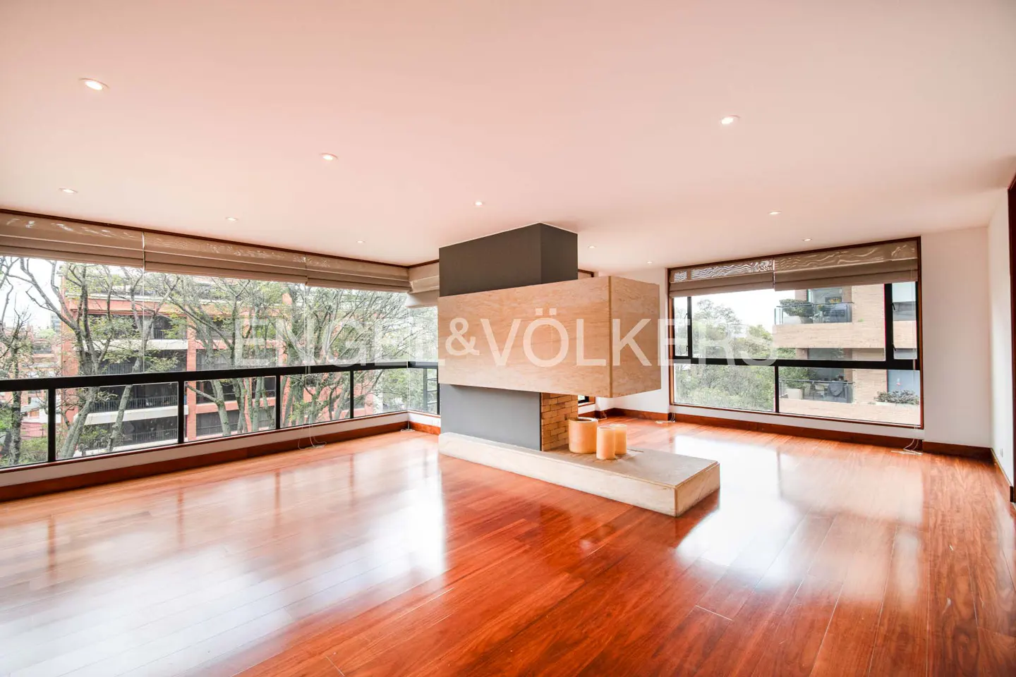 Bright, empty living room with hardwood floors, large windows, and a modern fireplace with candles. Trees visible outside.