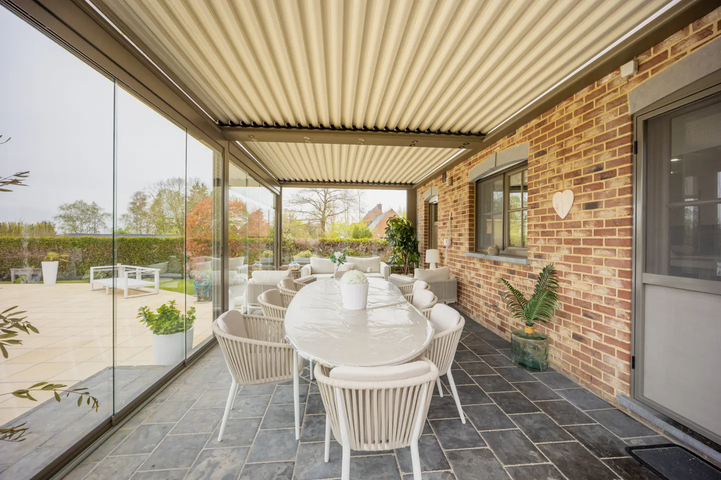 Covered patio with a long white table and chairs. Glass walls overlook a garden. Brick wall with window and door on the right.