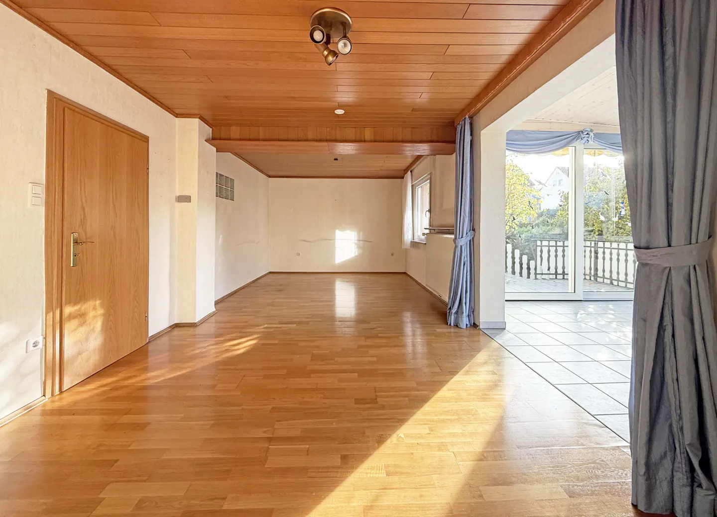 Bright, empty room with wood floors, a wood ceiling, and a door. A balcony is visible through an opening with blue curtains.