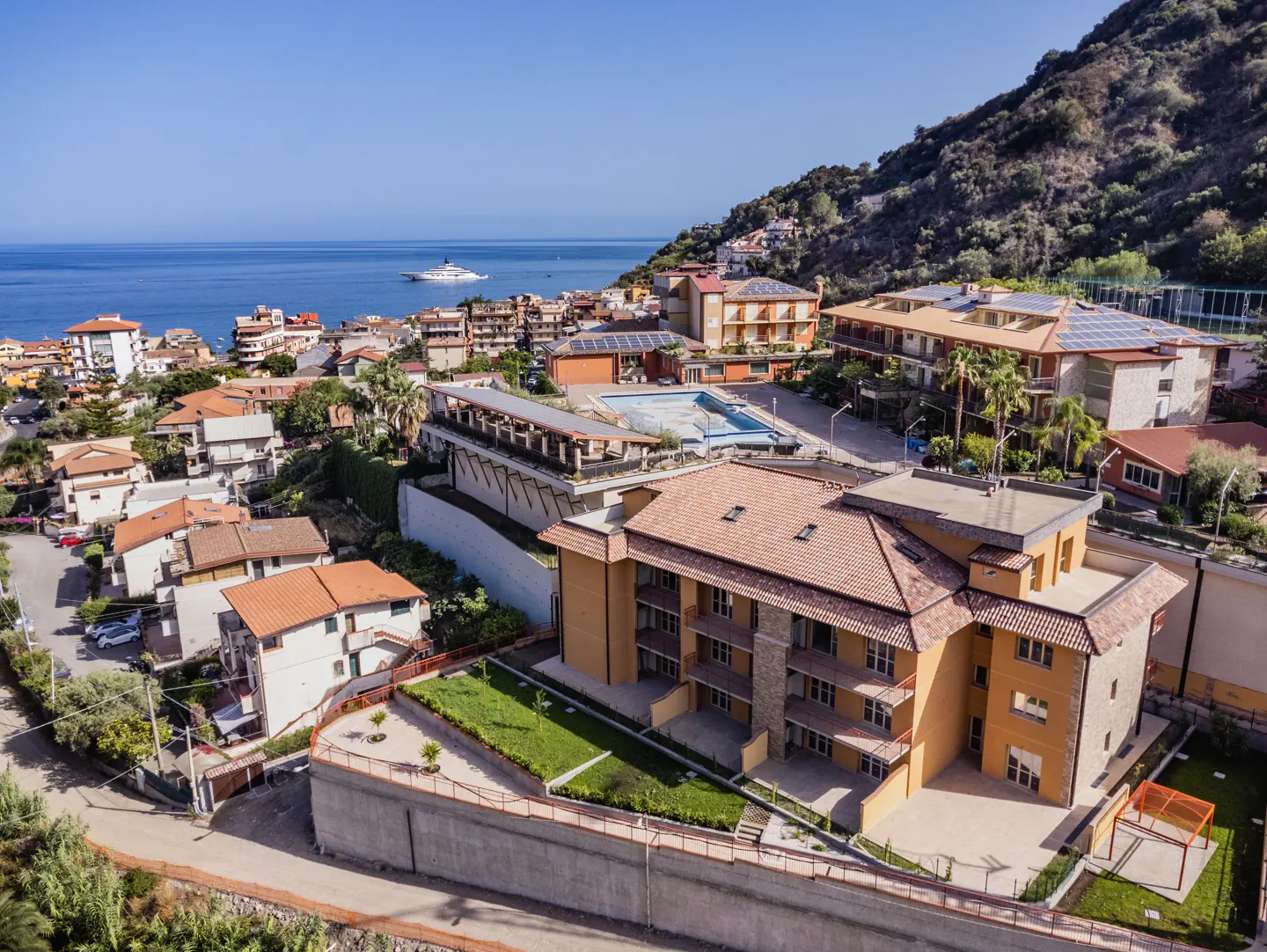 Aerial view of a Mediterranean resort with orange-roofed buildings, a swimming pool, and a hillside overlooking the sea.