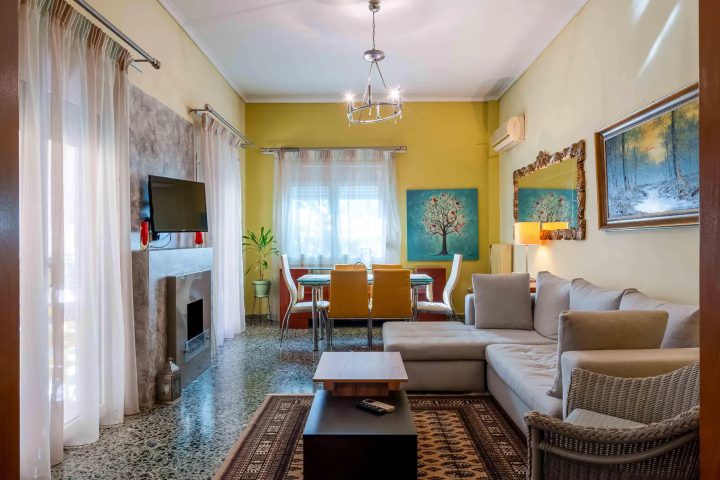 Living room with yellow walls, a gray sectional sofa, a dining table with orange chairs, and a TV above a fireplace.