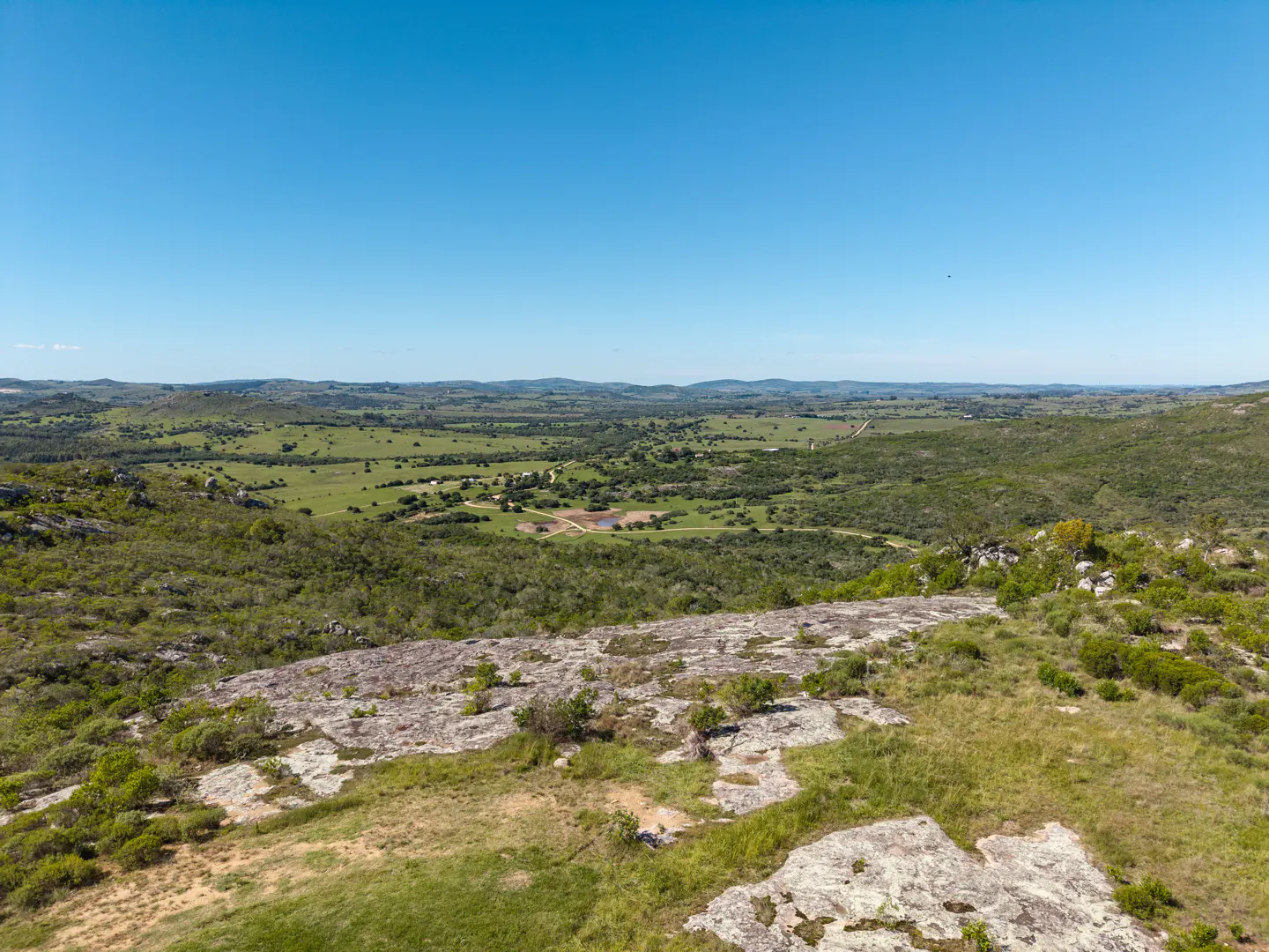 Scenic view of green fields and hills under a clear blue sky, seen from a rocky, grassy foreground.