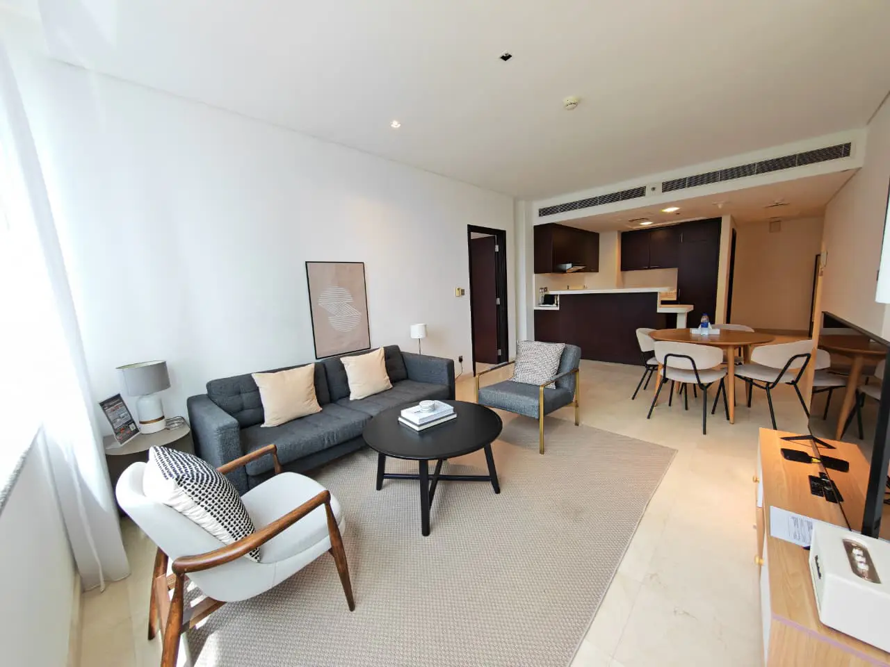A bright living room with a gray sofa, round black table, and open kitchen. A beige rug covers the floor.