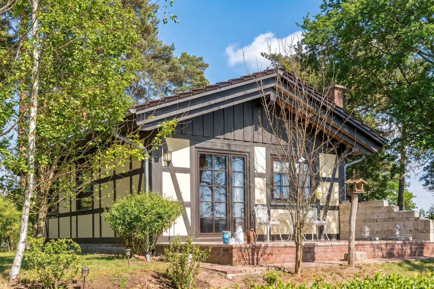 Charming half-timbered house with dark brown trim and a brick patio, surrounded by lush green trees under a bright blue sky.