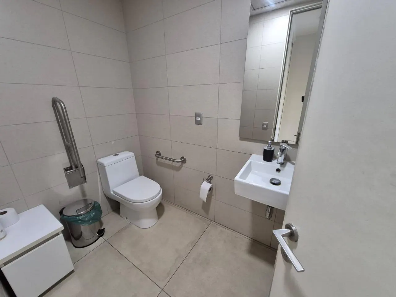 A modern bathroom with white toilet, sink, and tiled walls. Grab bars are installed for accessibility. A mirror hangs above the sink.