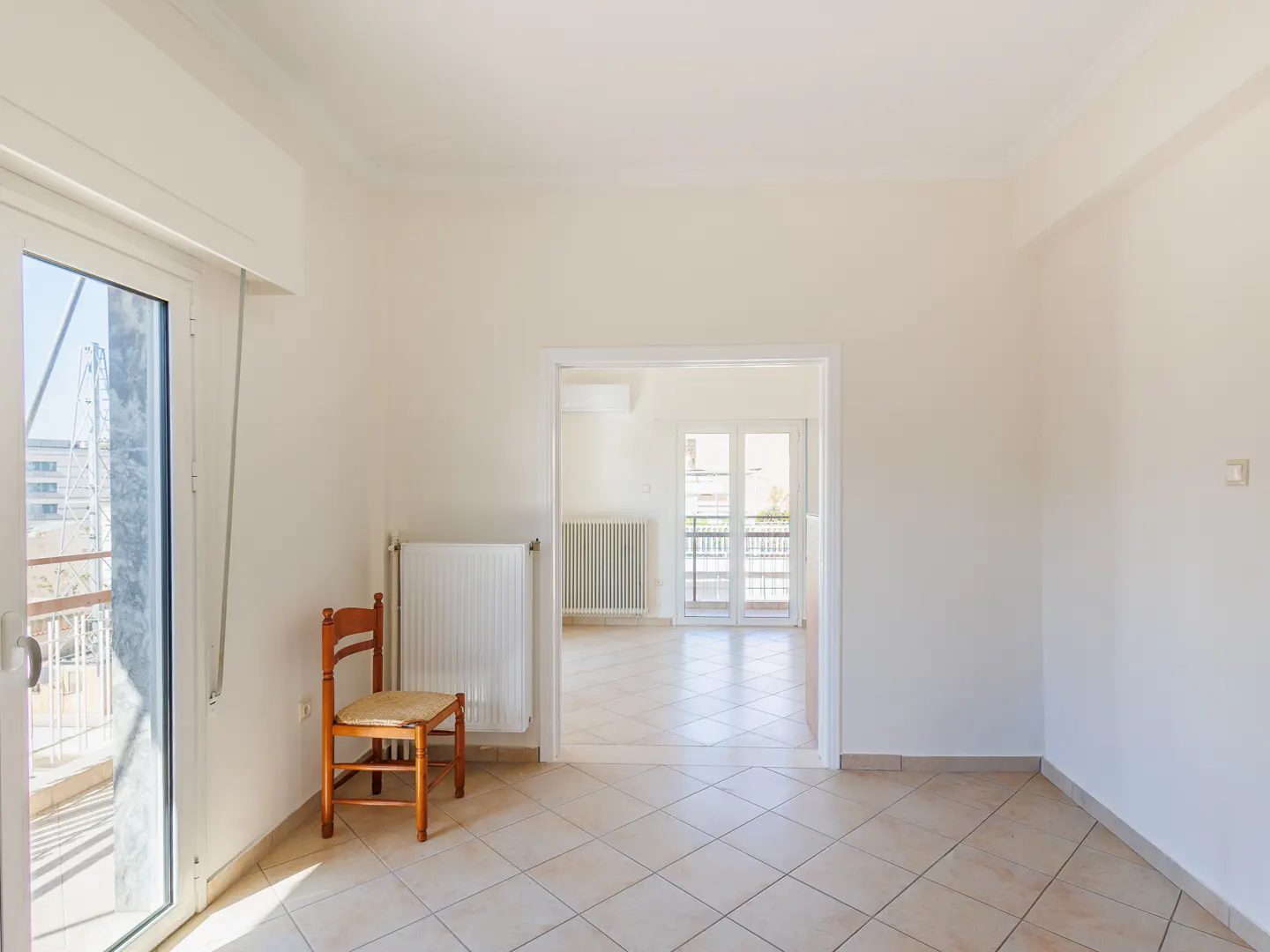 Bright, empty room with tile floor, white walls, and a wooden chair. A doorway leads to another room with a balcony. Natural light streams in.