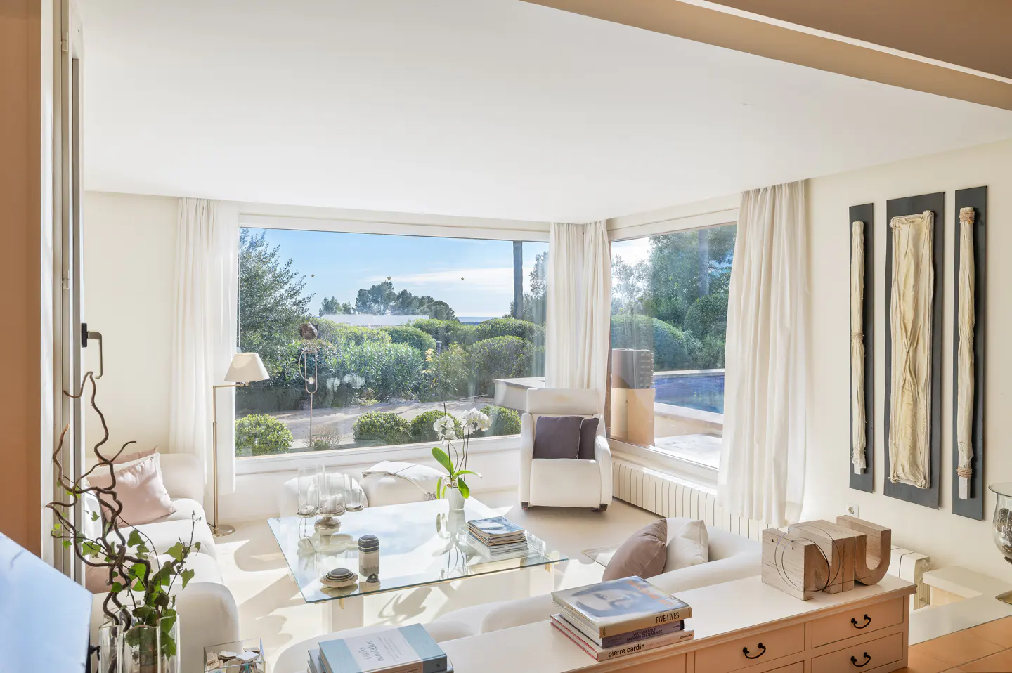 Bright living room with white furniture, a glass table, and large windows overlooking lush greenery and a pool. Modern art adorns the walls.