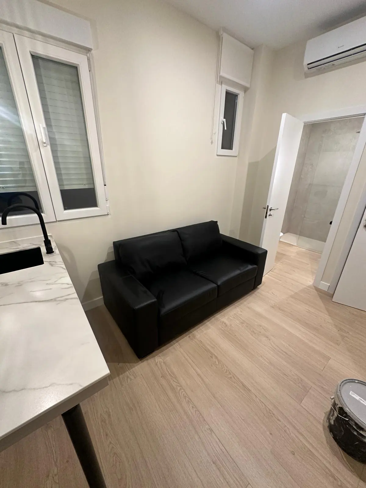 A bright, modern apartment interior with a black leather sofa, white walls, and light wood floors. A kitchen counter is visible on the left.