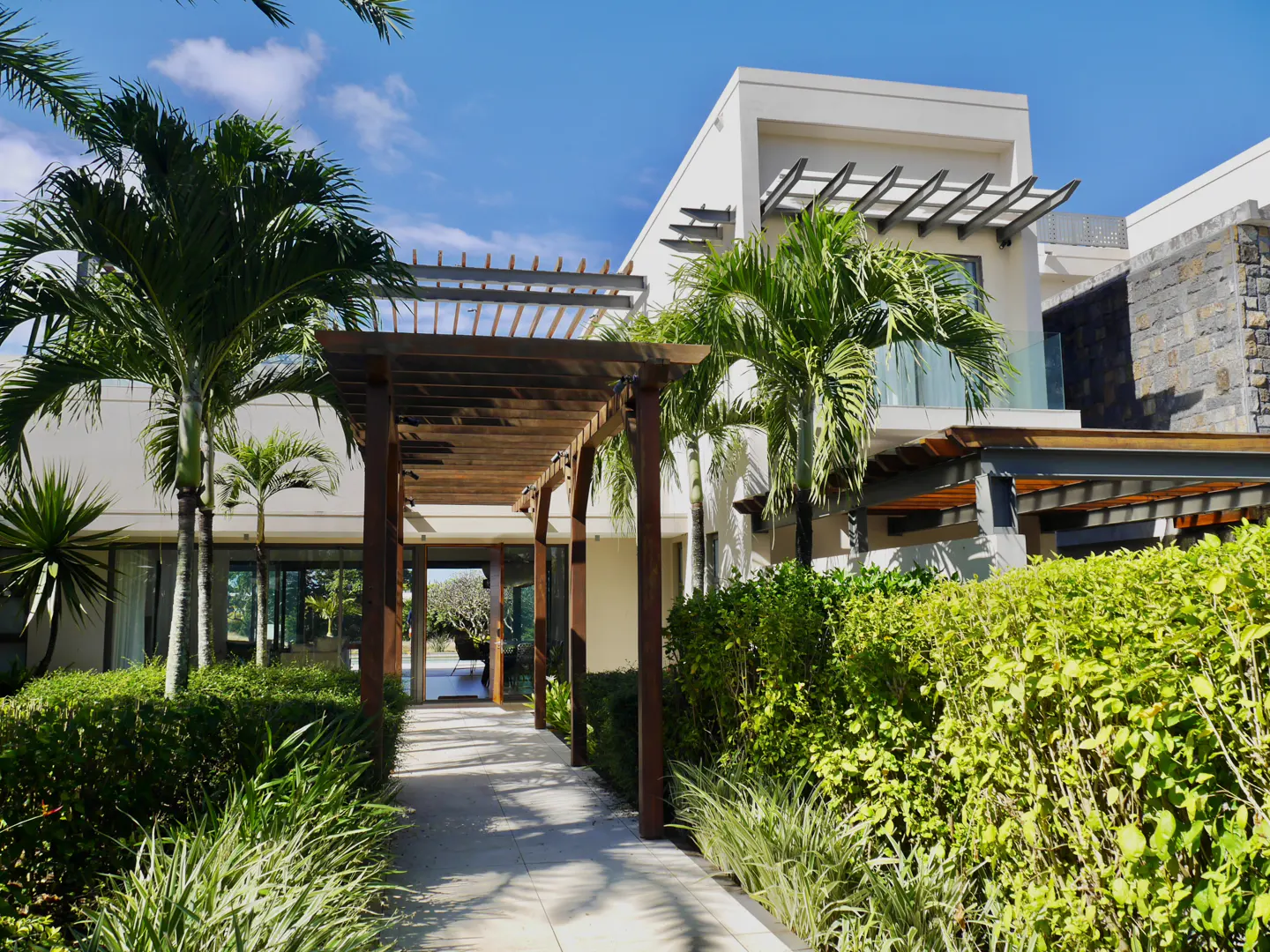 A modern white villa with a wooden pergola walkway, surrounded by lush green palm trees and manicured hedges under blue sky.