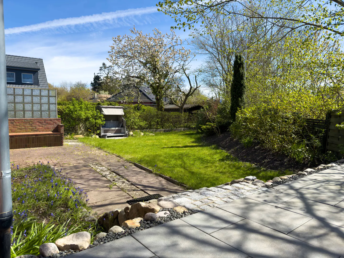 A backyard with a green lawn, trees, and a swing. A stone patio is in the foreground. The sky is blue with a few clouds.
