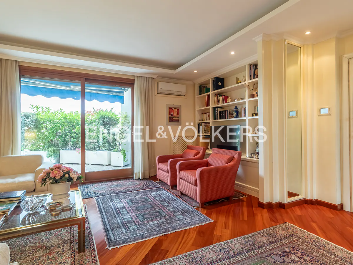 Bright living room with hardwood floors, two red armchairs, and a bookshelf. A glass door leads to a balcony with plants.