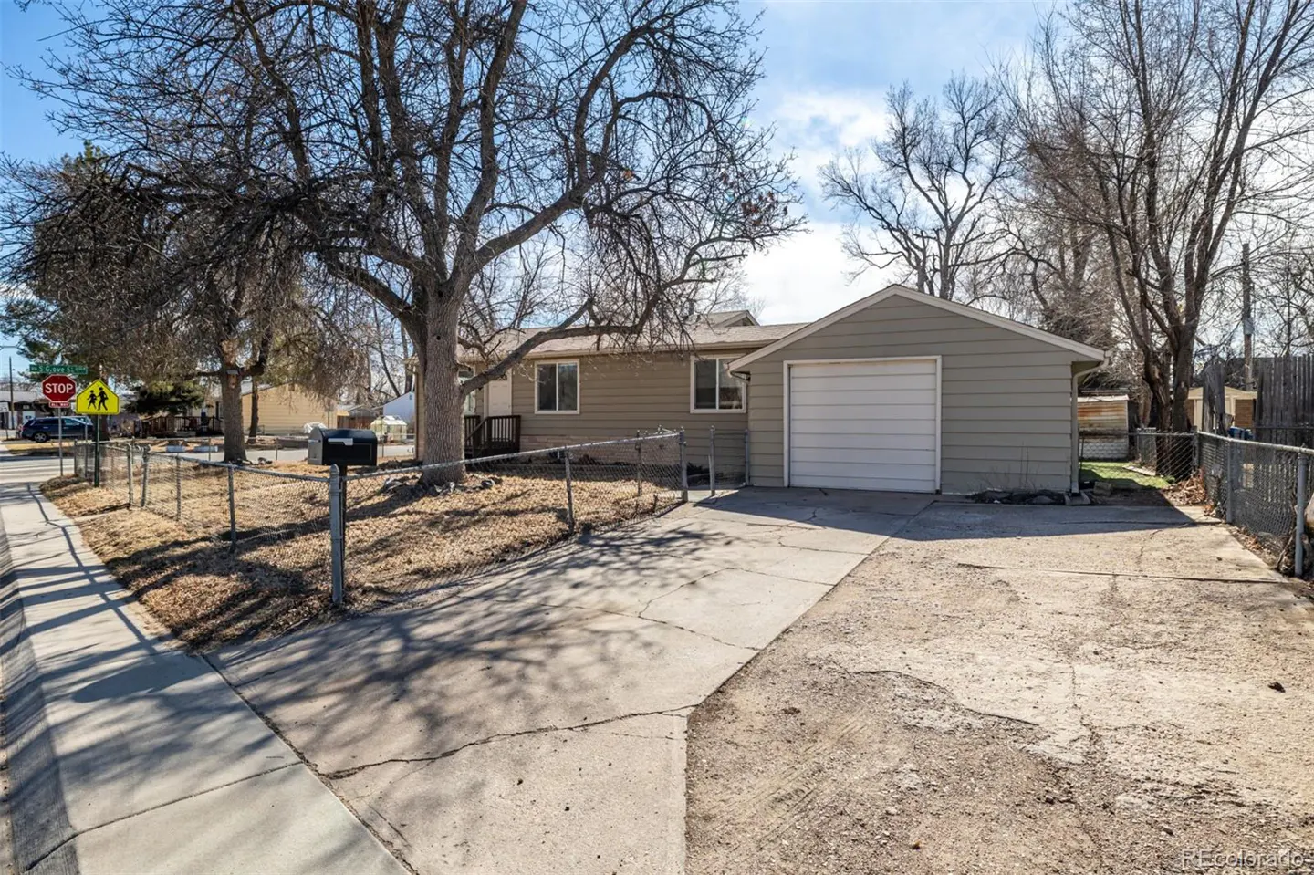 A single-story beige house with a white garage door, a chain-link fence, and bare trees in the front yard.