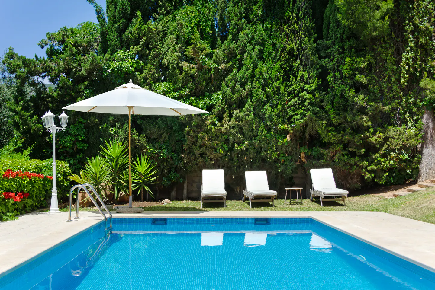 A blue swimming pool with three white lounge chairs, a white umbrella, and a lamp post against a backdrop of lush trees.