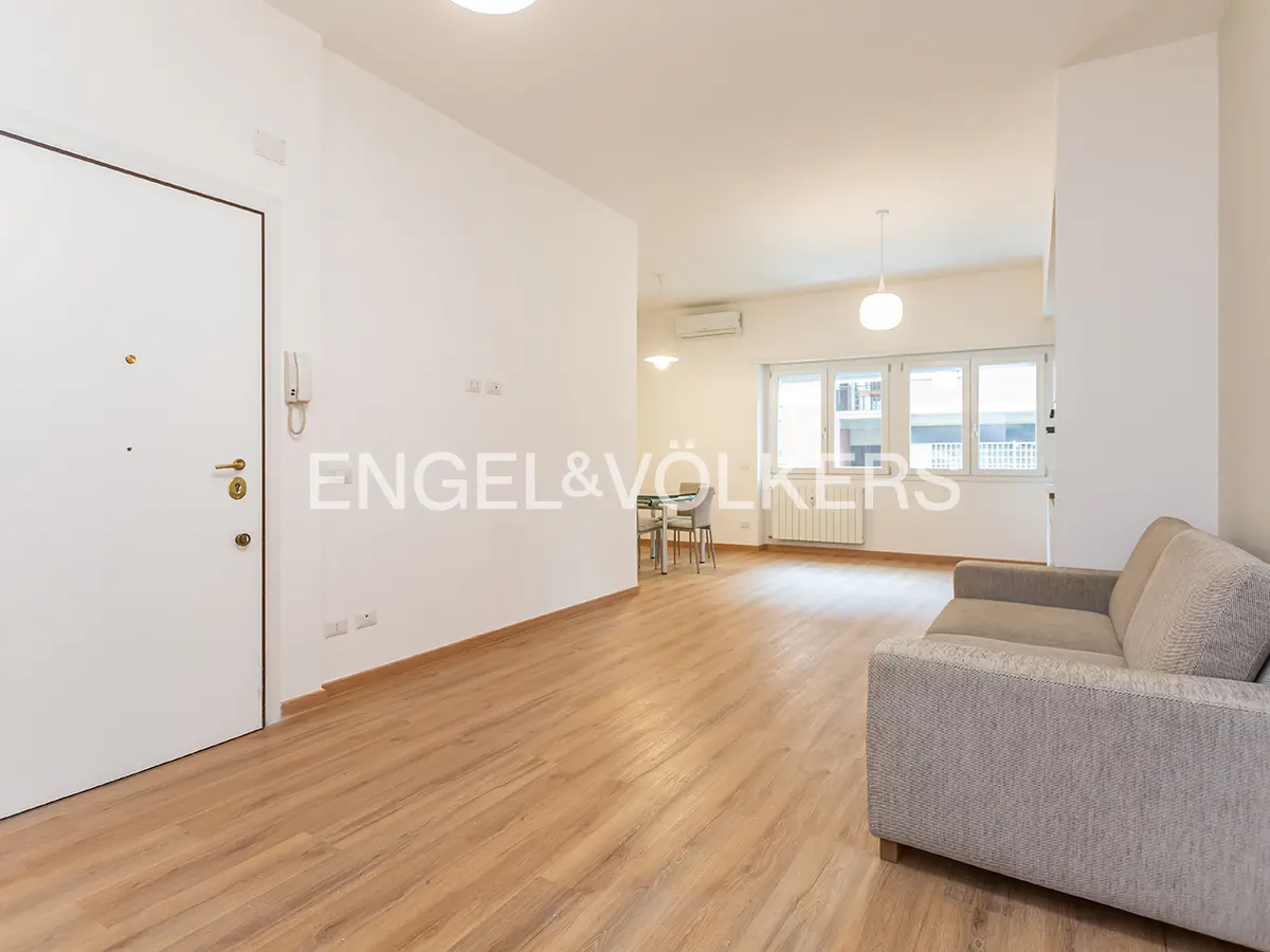 Bright, modern apartment interior with light wood floors, white walls, and a gray sofa. Dining table and windows in the background.