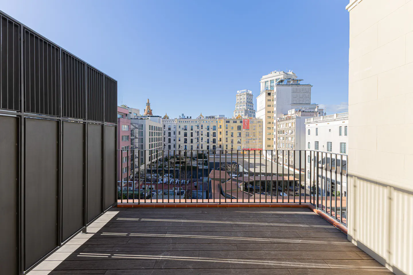 Balcony view with dark wood floor, black metal railings, and city buildings in the background under a clear blue sky.