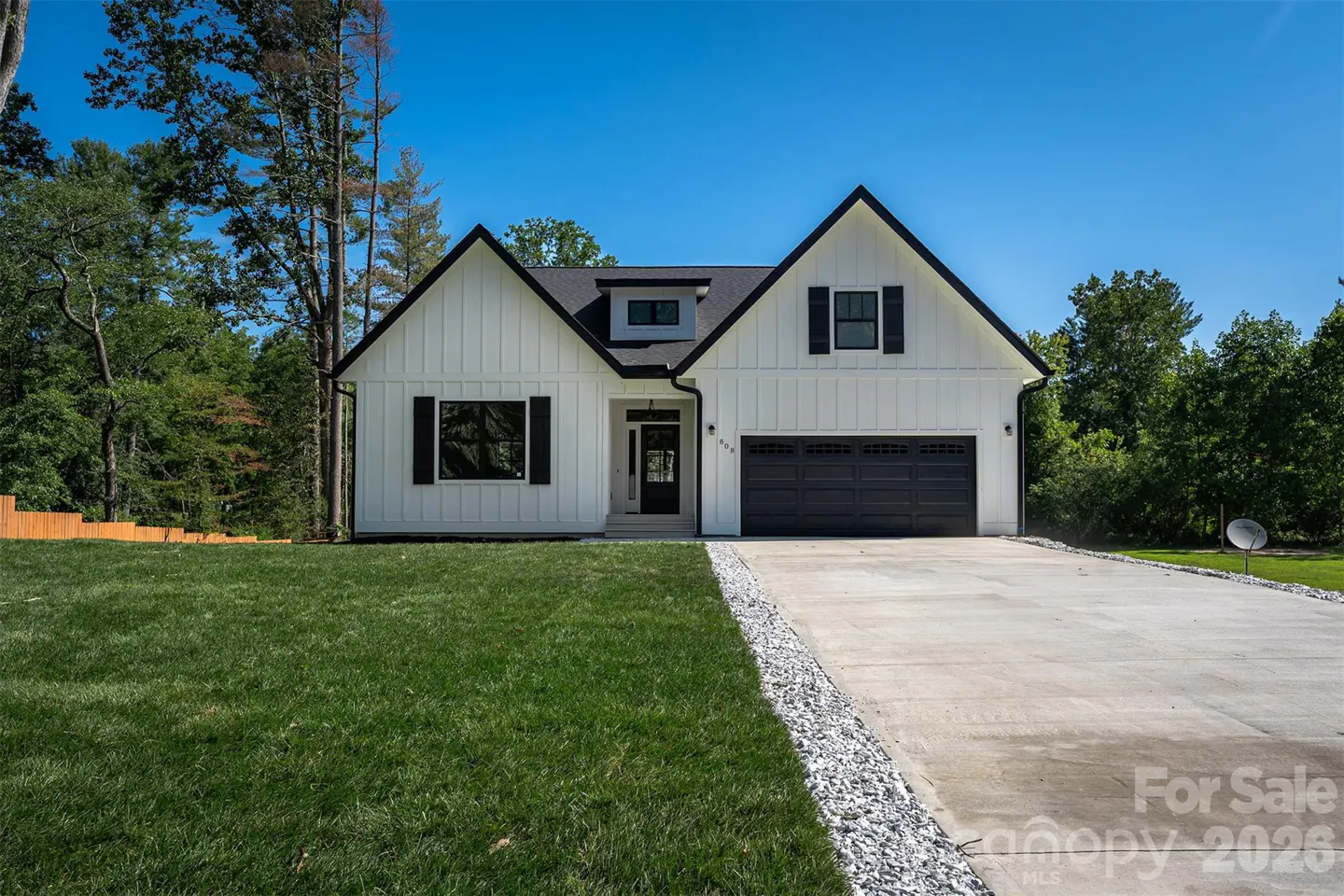 A modern farmhouse with white siding, black trim, and a dark gray garage door sits on a green lawn under a blue sky.