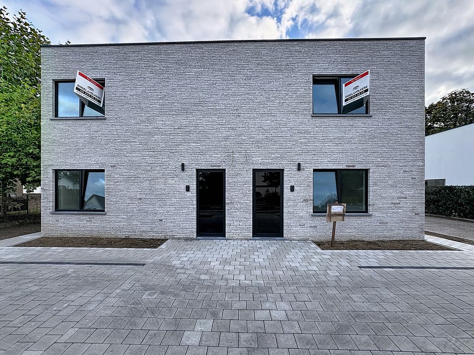 Modern two-story gray brick duplex with black doors and window frames, featuring "For Sale" signs in the upper windows. [AI-generated text]