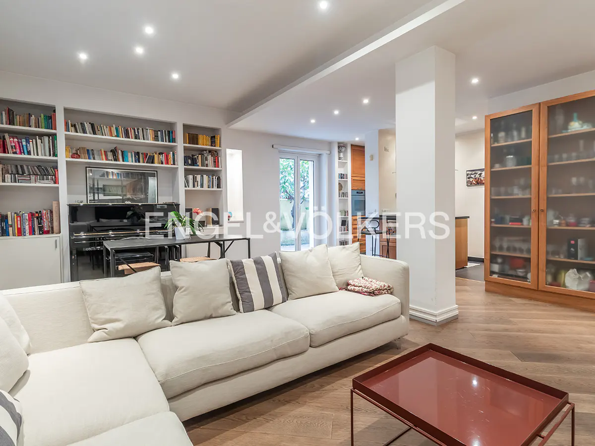 Bright living room with white sectional sofa, bookshelves, piano, and wood floors. A red coffee table sits in the foreground.