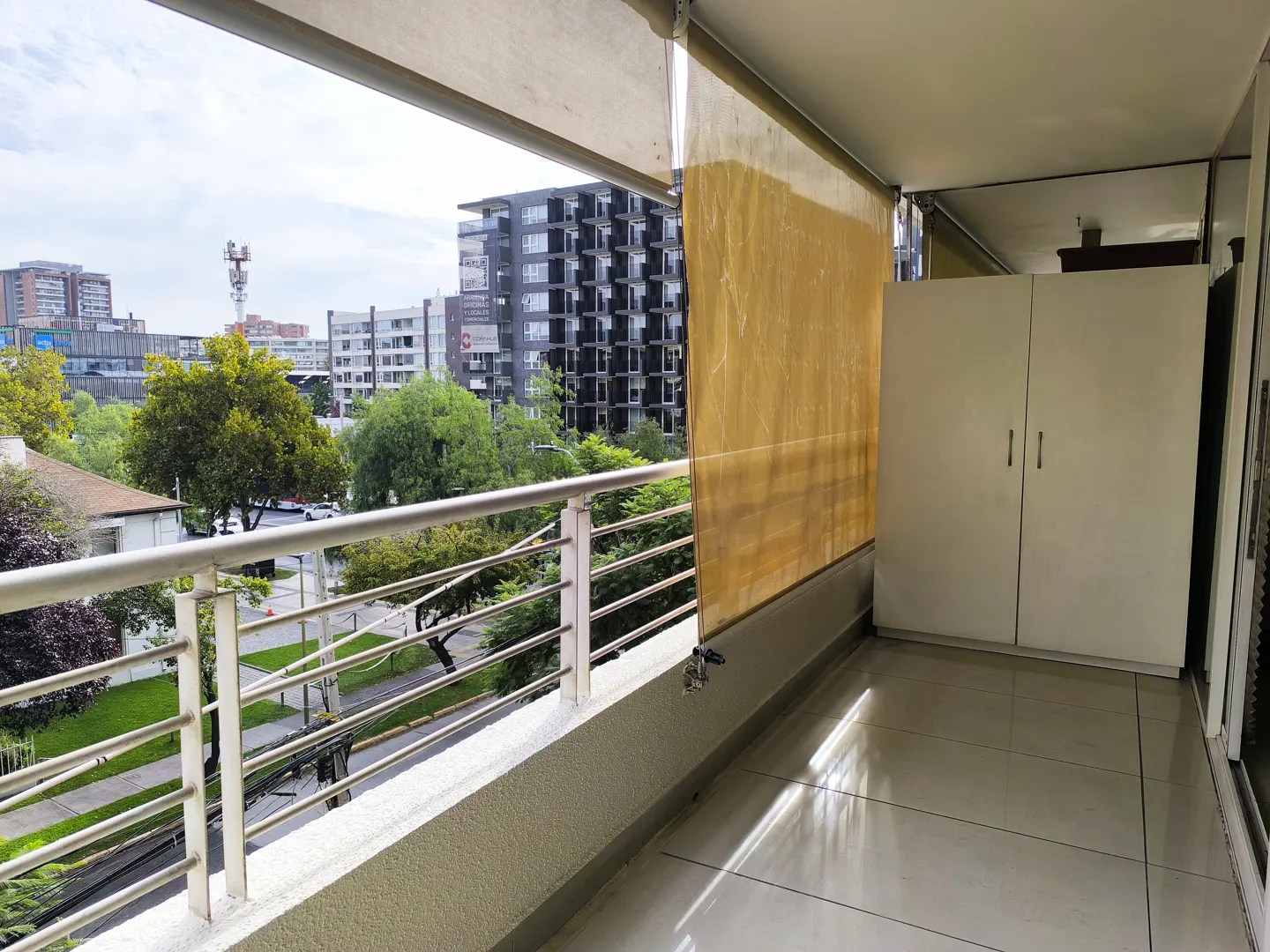 Balcony view with metal railing, tan sunshade, and white storage cabinet. City buildings and green trees in the background.