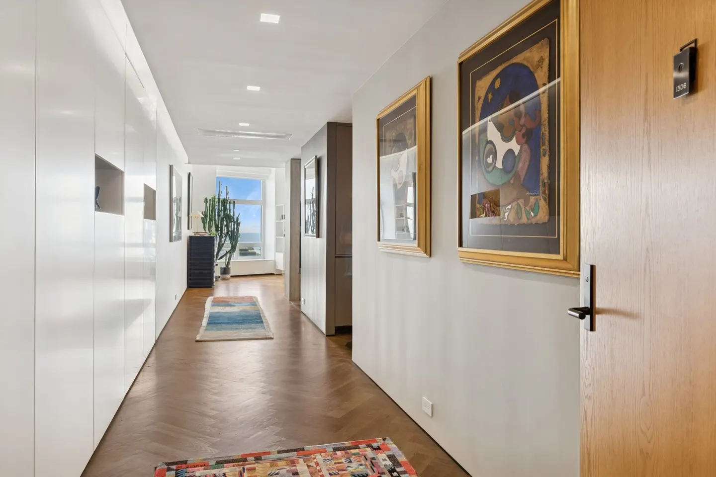 Hallway with herringbone wood floors, white walls, and art. A cactus sits near a window with an ocean view.