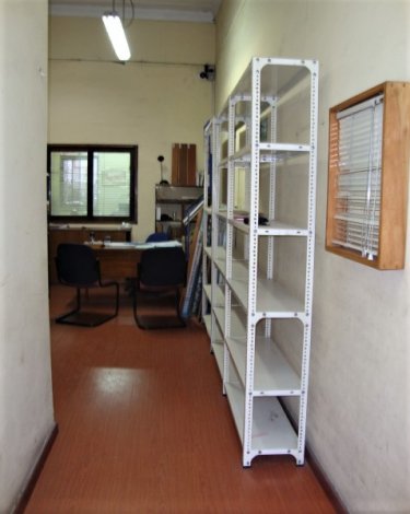 A narrow hallway with a red floor leads to an office with a desk, chairs, and window. White metal shelving lines the right wall.