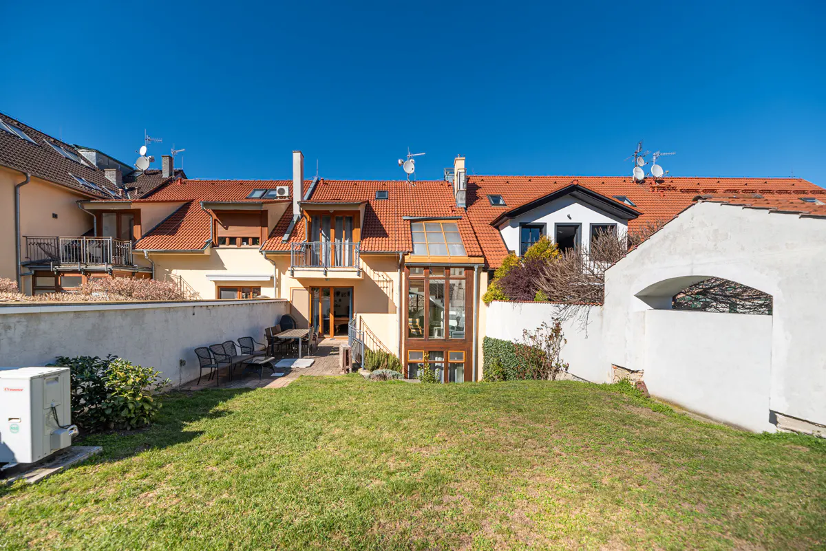 Backyard view of a yellow townhouse with a red tile roof, a green lawn, and a white wall under a clear blue sky.