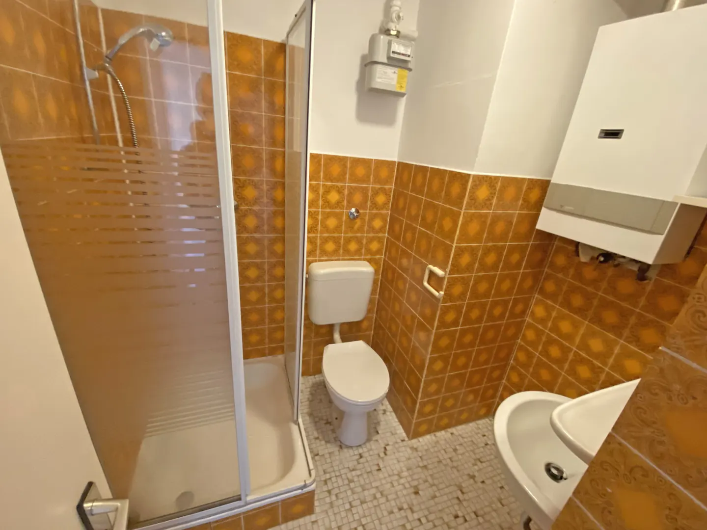 Bathroom with orange patterned wall tiles, a glass shower stall, white toilet, sink, and a wall-mounted water heater.