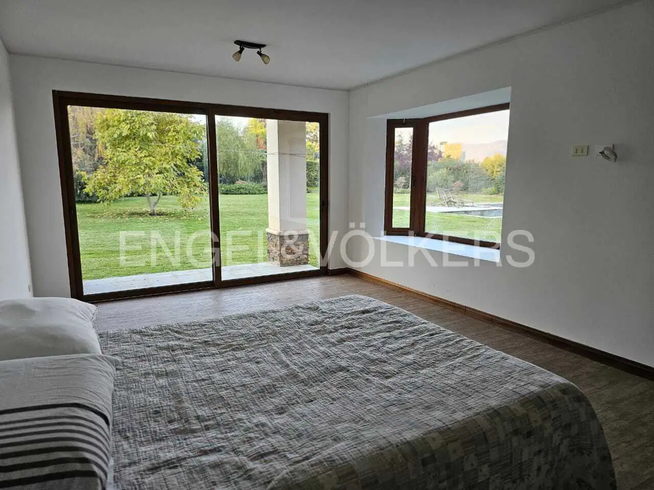 Bedroom with a gray patterned bedspread, large windows overlooking a green lawn and trees. The walls are white.