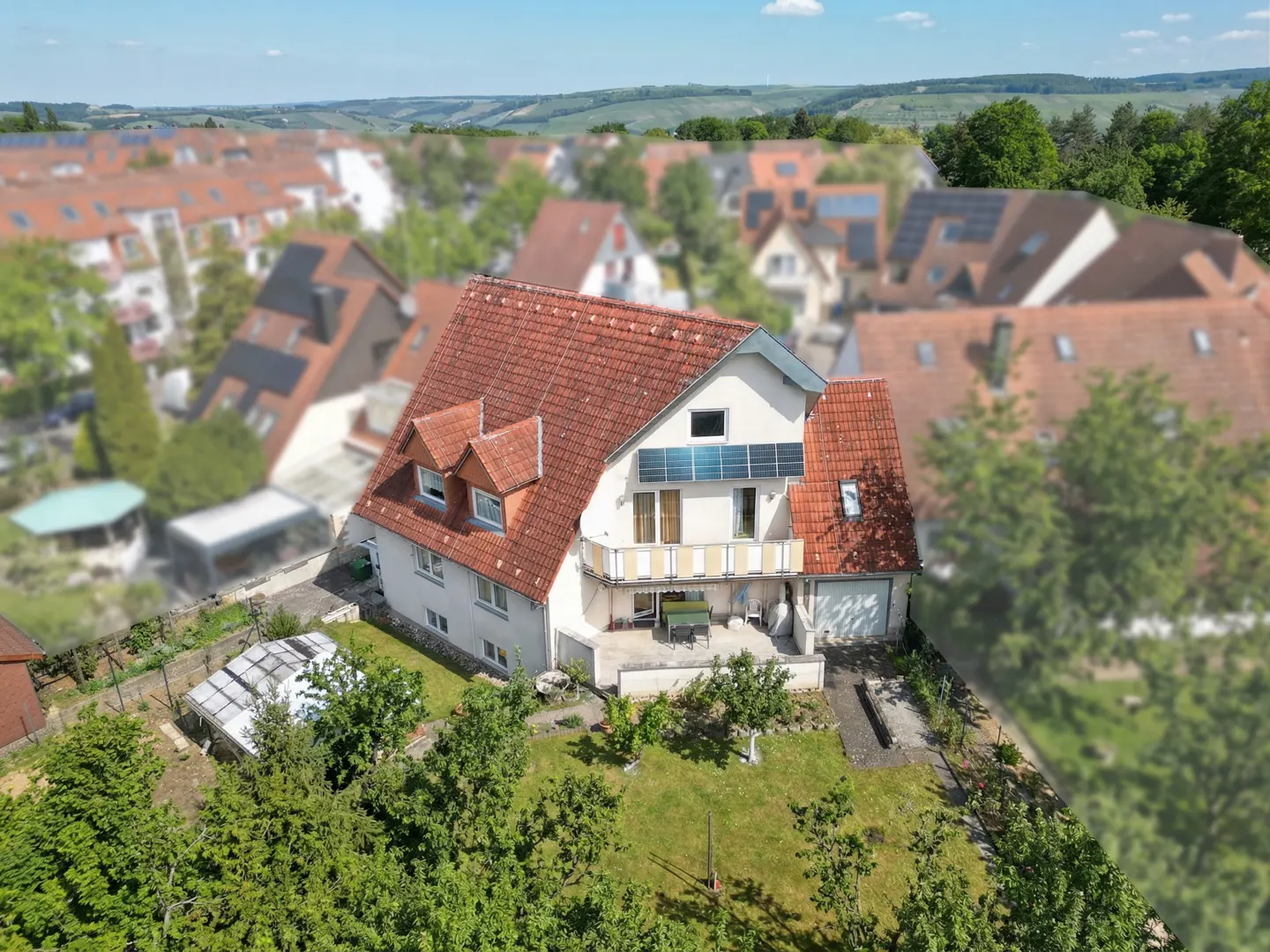 Aerial view of a two-story white house with a red tile roof and solar panels, surrounded by green trees and other houses.