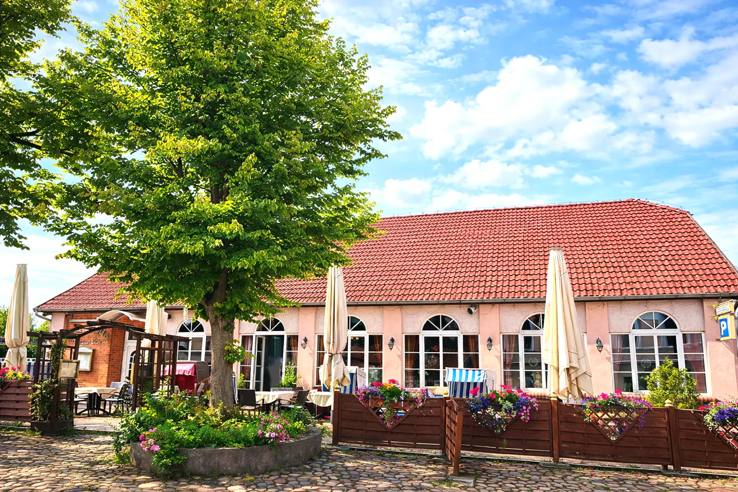 Outdoor patio of a pink building with a red tile roof, large tree, and tables under umbrellas on a sunny day.