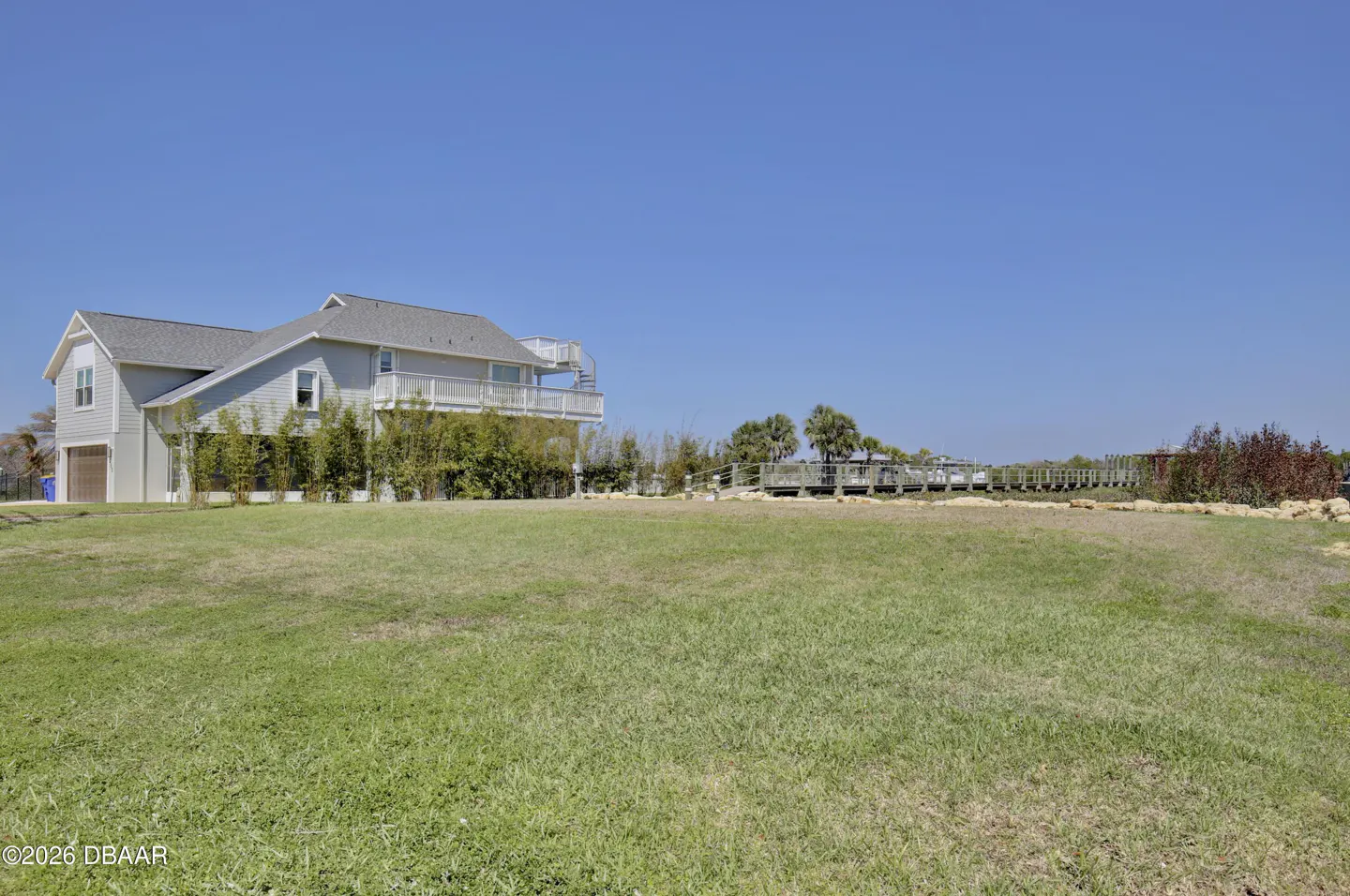 A two-story gray house with a deck and spiral staircase, viewed across a large green lawn under a clear blue sky.