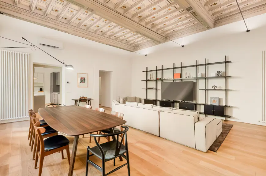 Bright, open living space with wood floors, a long dining table, and a large white sofa facing a TV on a black shelving unit.