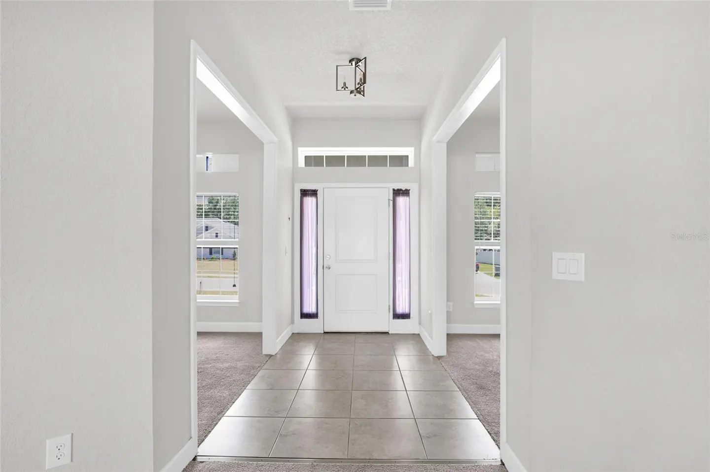 A bright foyer with gray tile floors leads to a white front door with sidelights. Two open doorways lead to carpeted rooms with windows.
