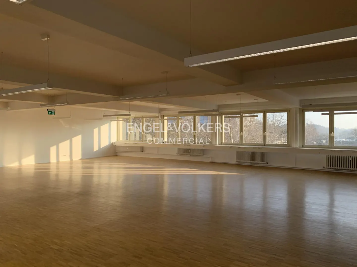 Empty commercial space with hardwood floors, white walls, and large windows. Sunlight streams in, illuminating the room.