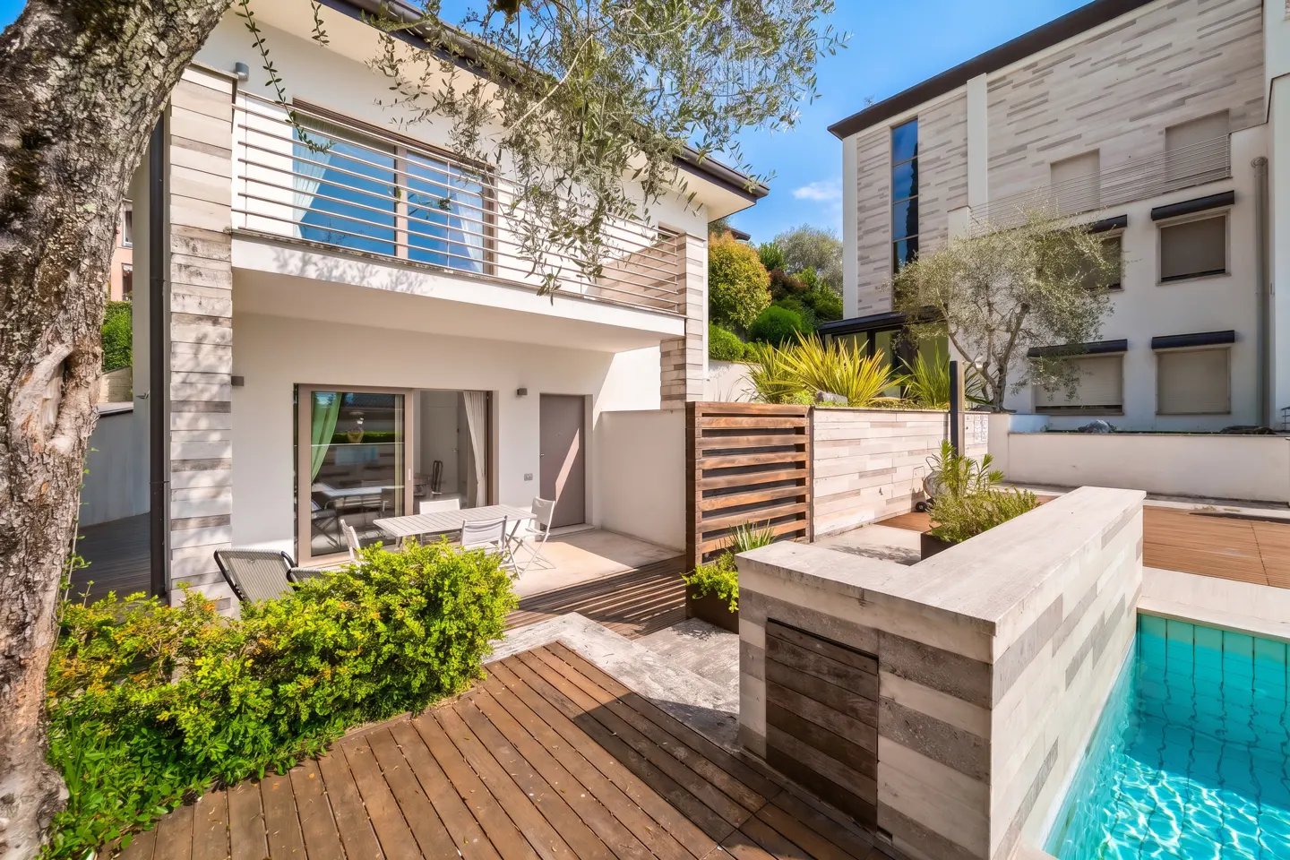 Exterior view of a modern two-story house with a wooden deck, a pool, and outdoor furniture.