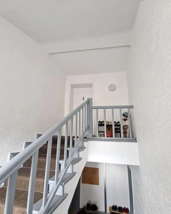 Interior view of a white stairwell with gray railings, brown carpeted stairs, and a shoe rack on the landing.