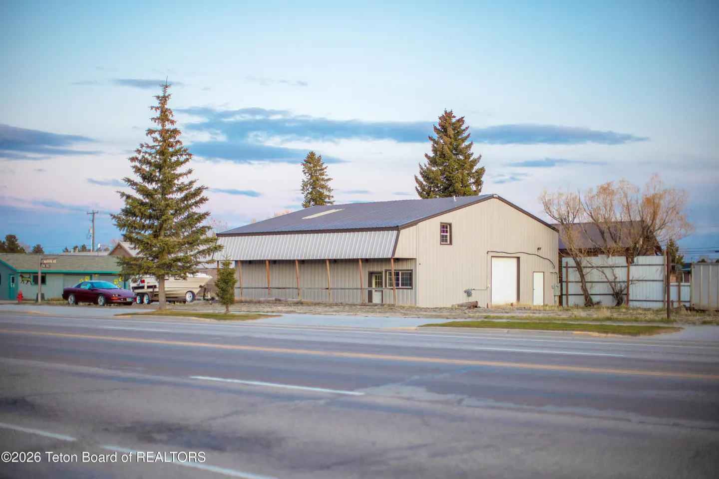 A gray metal building with a dark roof sits near a road, with trees and a car visible.