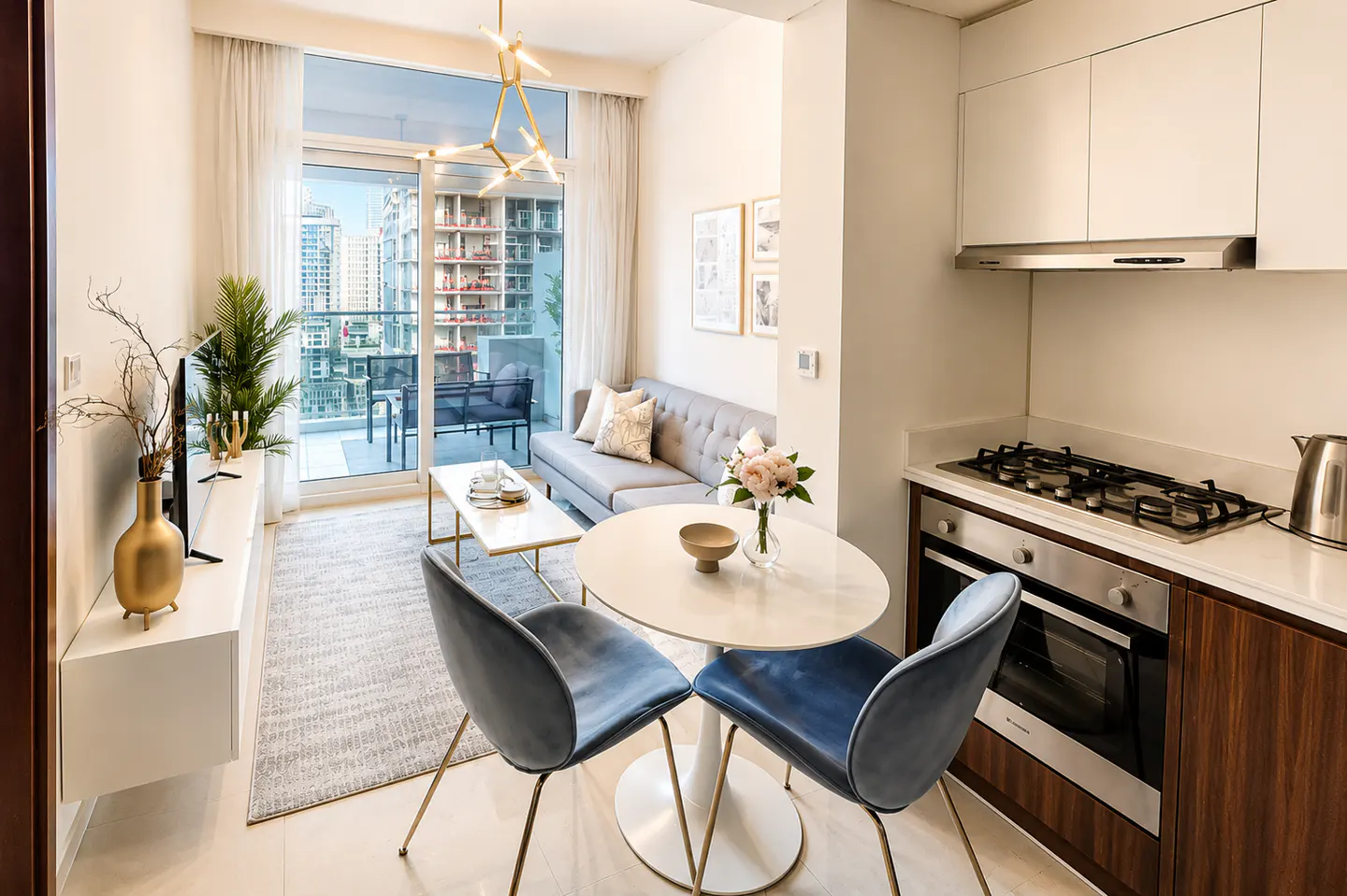 Bright apartment interior with a gray sofa, round table with two blue chairs, and a balcony overlooking city buildings.