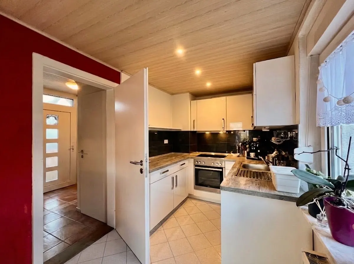A bright kitchen with white cabinets, dark countertops, and a window with plants. A doorway leads to a red-walled hallway with a front door.