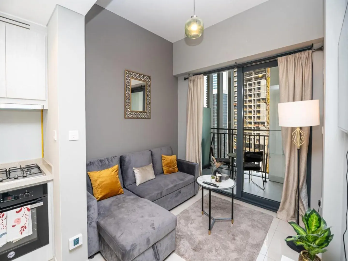 Living room with gray sofa, yellow pillows, and balcony view of city buildings. Kitchenette visible on the left.