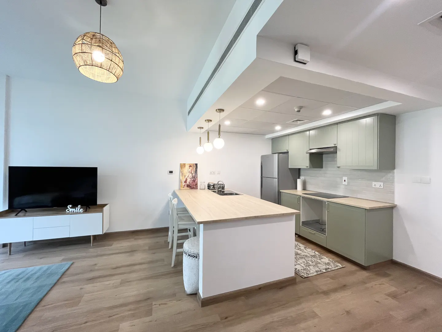A modern apartment interior with a kitchen island, sage green cabinets, and a living area with a TV.
