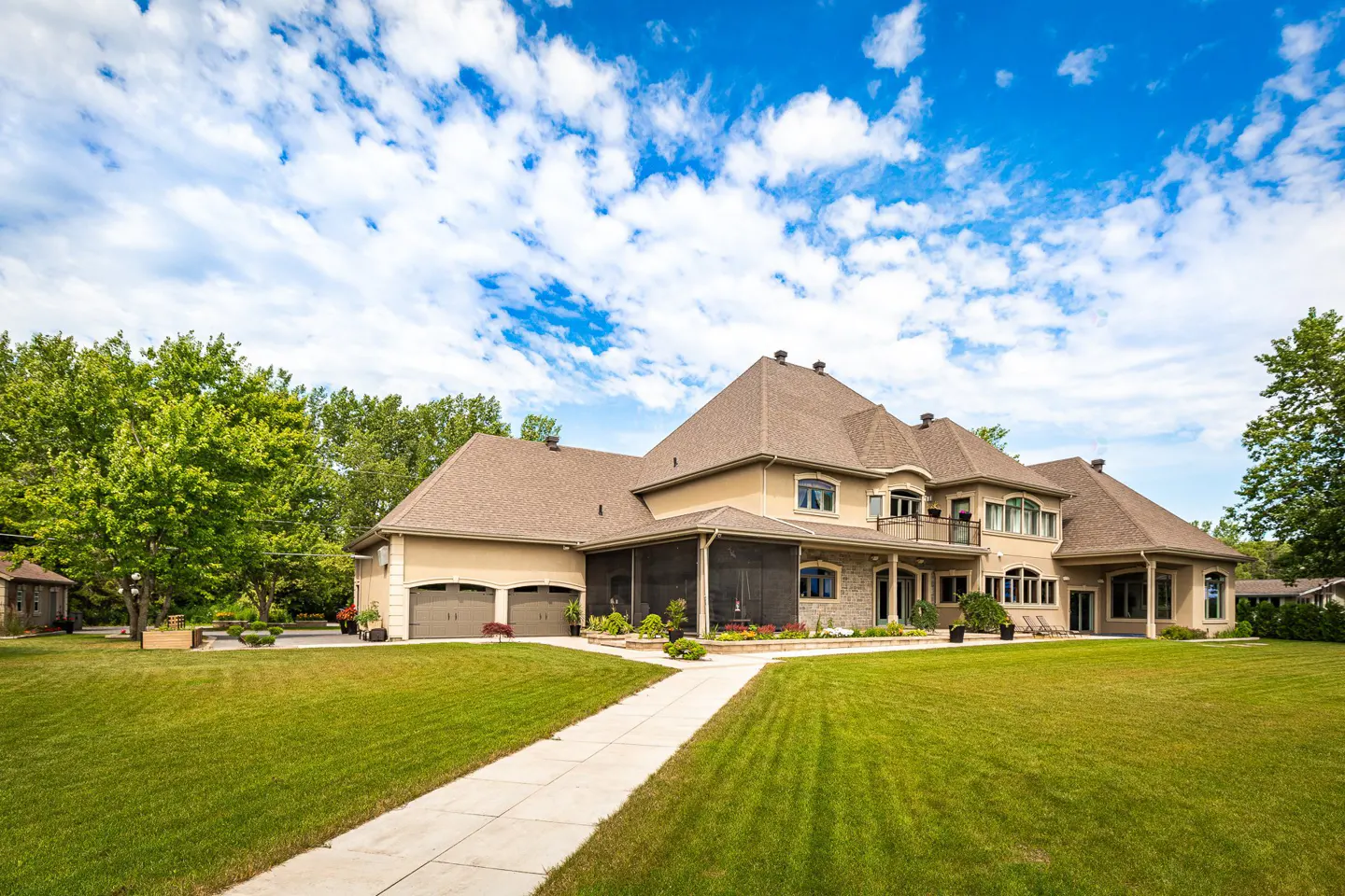 Beige two-story house with a brown roof, a two-car garage, and a large green lawn under a blue sky with white clouds.