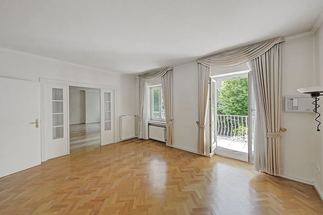 Bright, empty room with herringbone wood floors, white walls, and balcony access with beige curtains.
