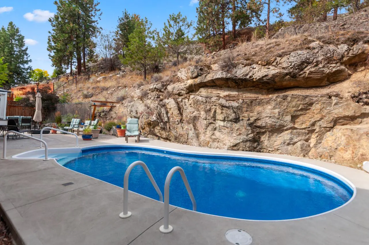 Backyard pool with blue water and white ladder, surrounded by a concrete patio. A rocky hill rises in the background.
