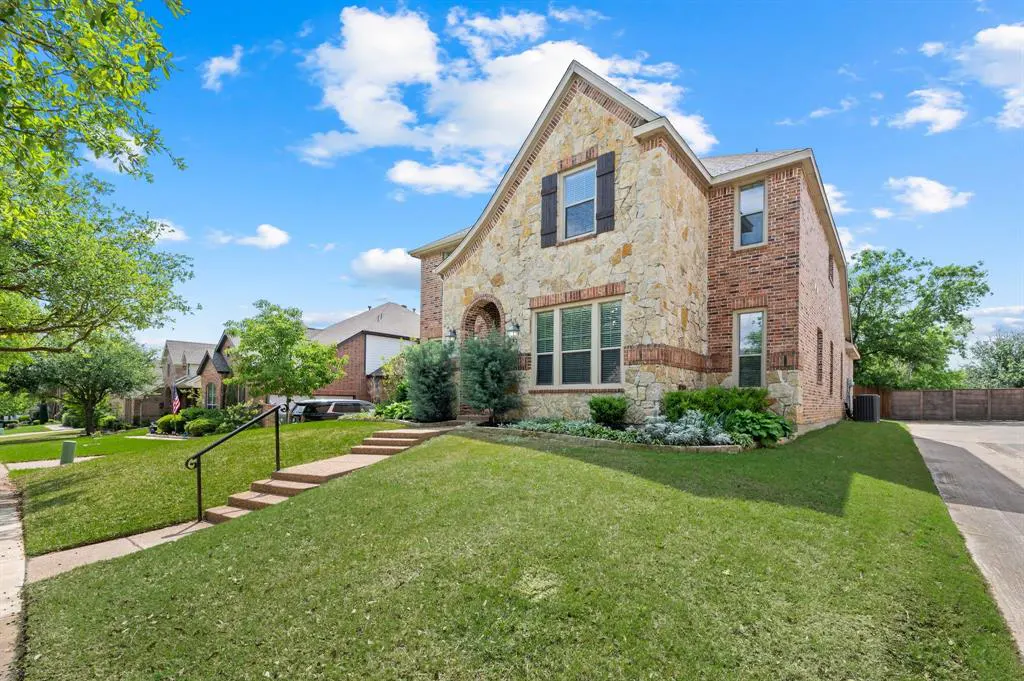 Two-story house with stone and brick exterior, dark shutters, green lawn, and blue sky with clouds.