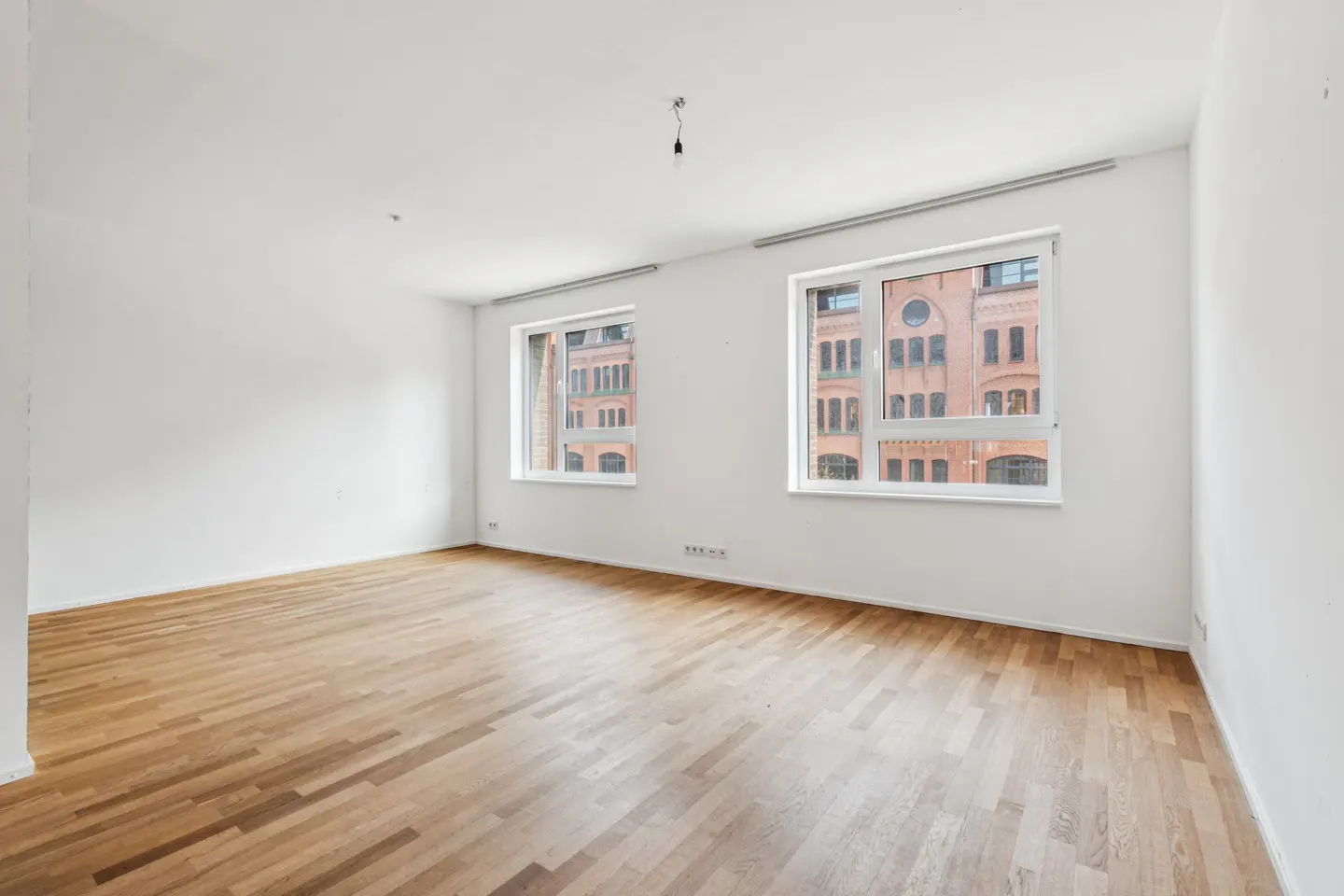 Bright, empty room with hardwood floors and white walls. Two windows show a brick building outside.