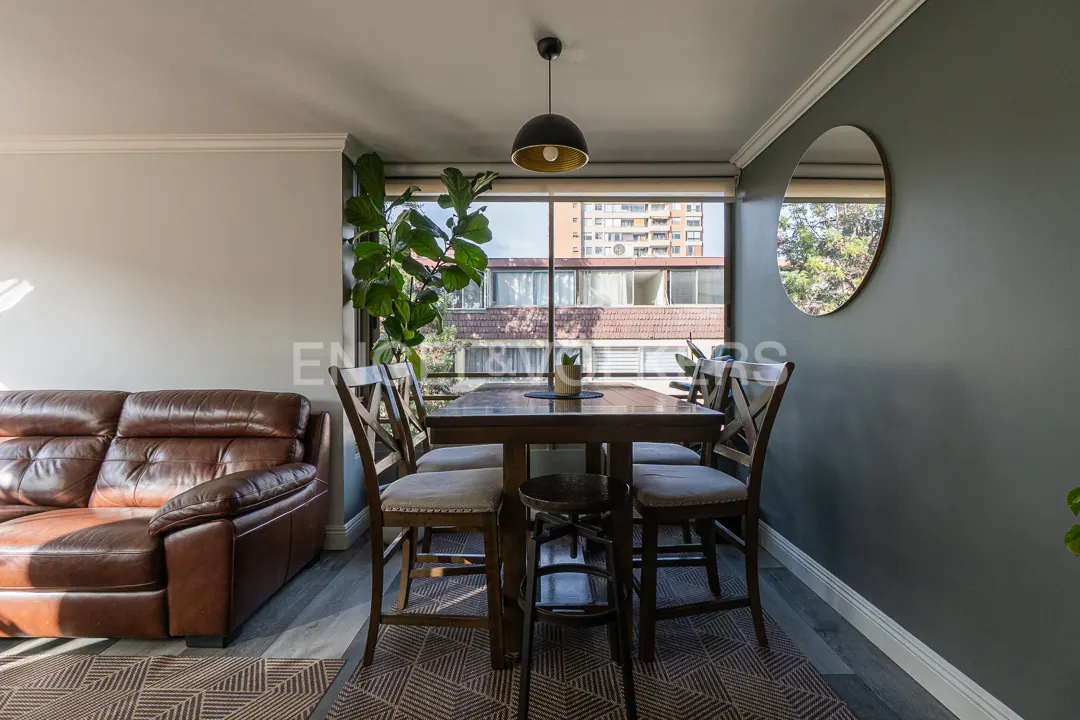 Dining area with wooden table, four chairs, and a stool, next to a brown leather sofa, a large plant, and a round mirror.