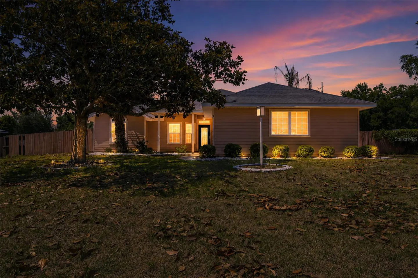 Exterior shot of a tan single-story home at dusk with a large tree in the front yard. Windows glow with warm light.