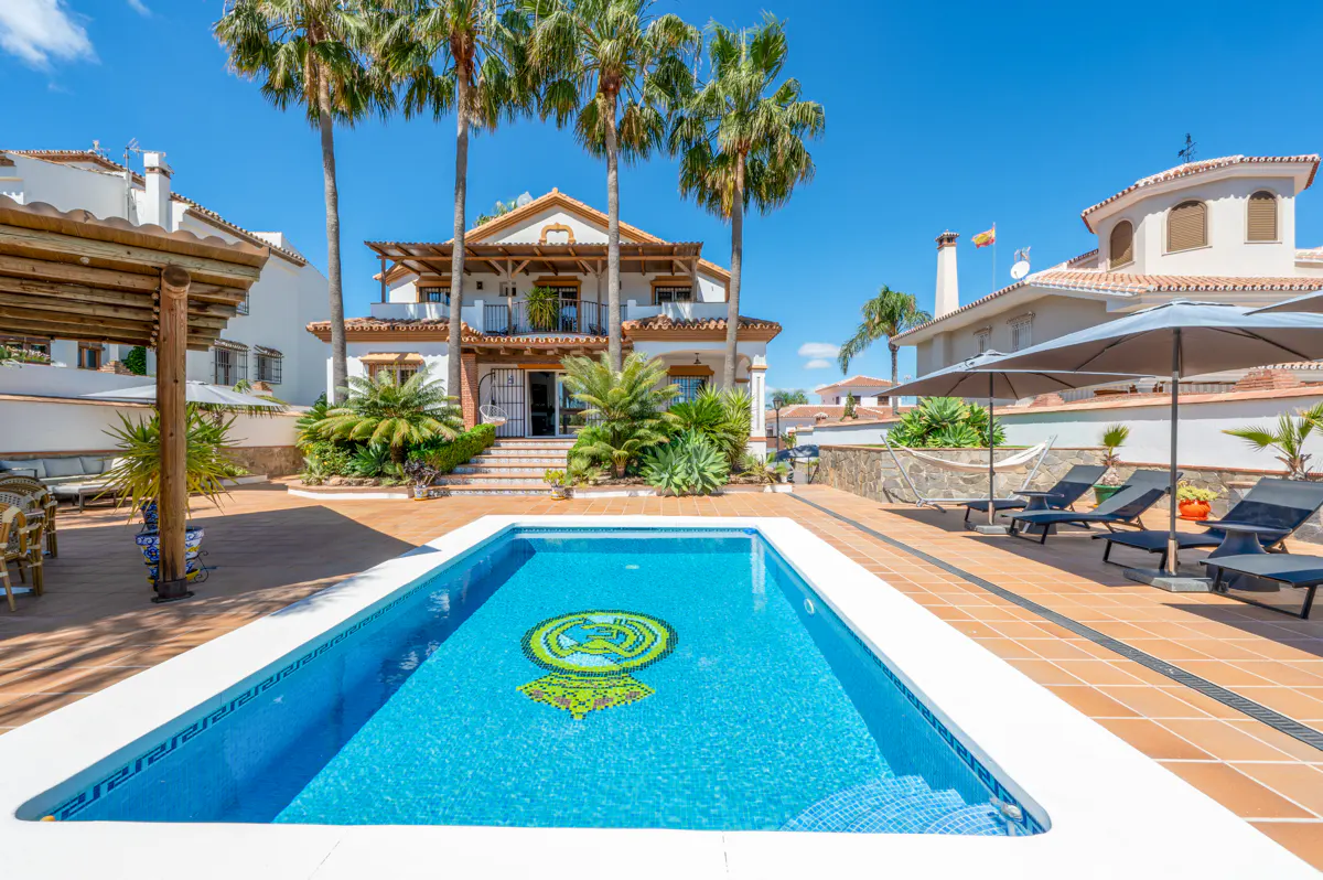 A bright blue pool with a mosaic design sits in front of a white two-story house with palm trees. Lounge chairs and umbrellas are nearby.